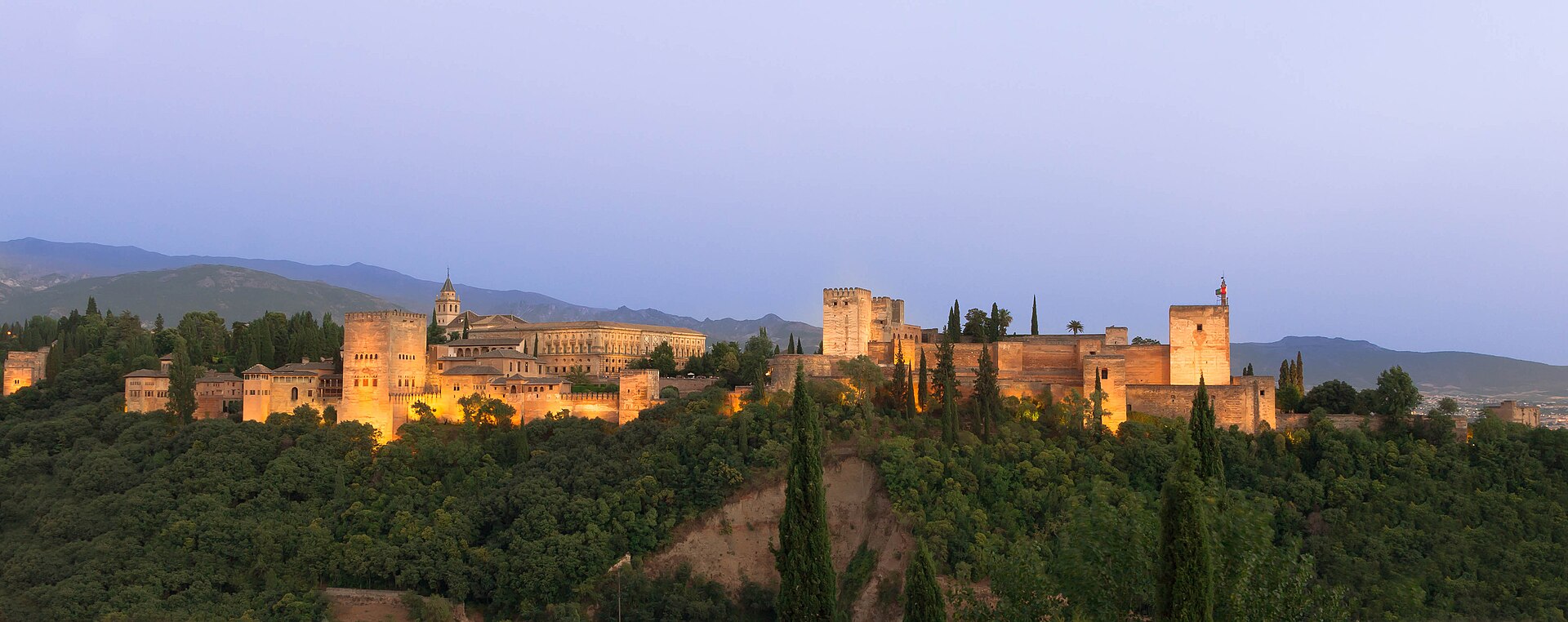 Vista panorámica al atardecer de la Alhambra de Granada sobre la colina arbolada, con Sierra Nevada al fondo