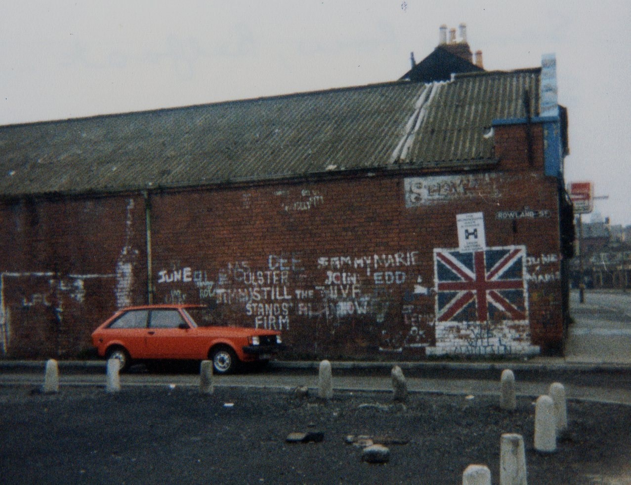 Fotografía en color de una nave de ladrillo con pintadas, una bandera británica pintada en la pared y un coche rojo aparcado delante en una calle vacía