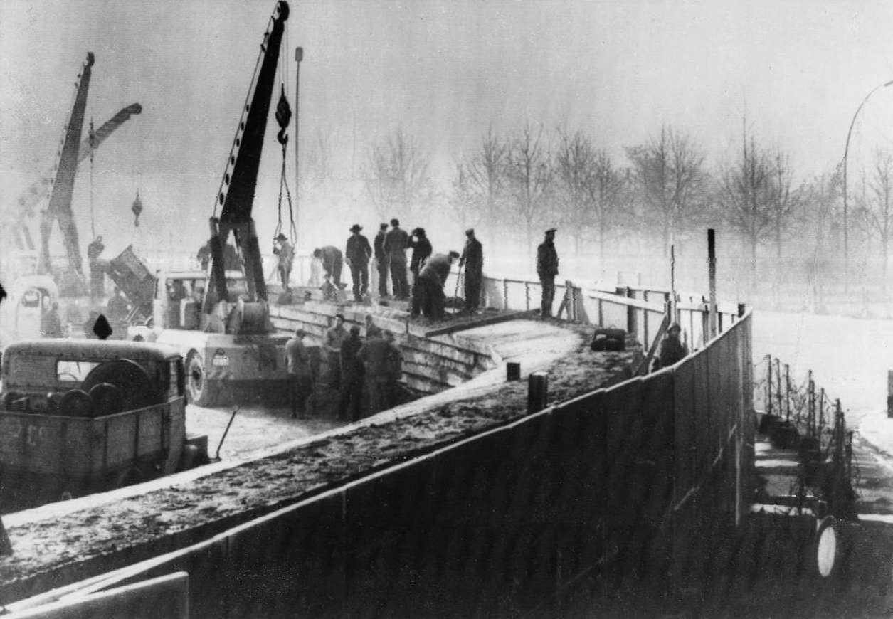 Fotografía en blanco y negro de un muro de bloques en construcción con grúas y trabajadores, y un grupo de personas observando al otro lado