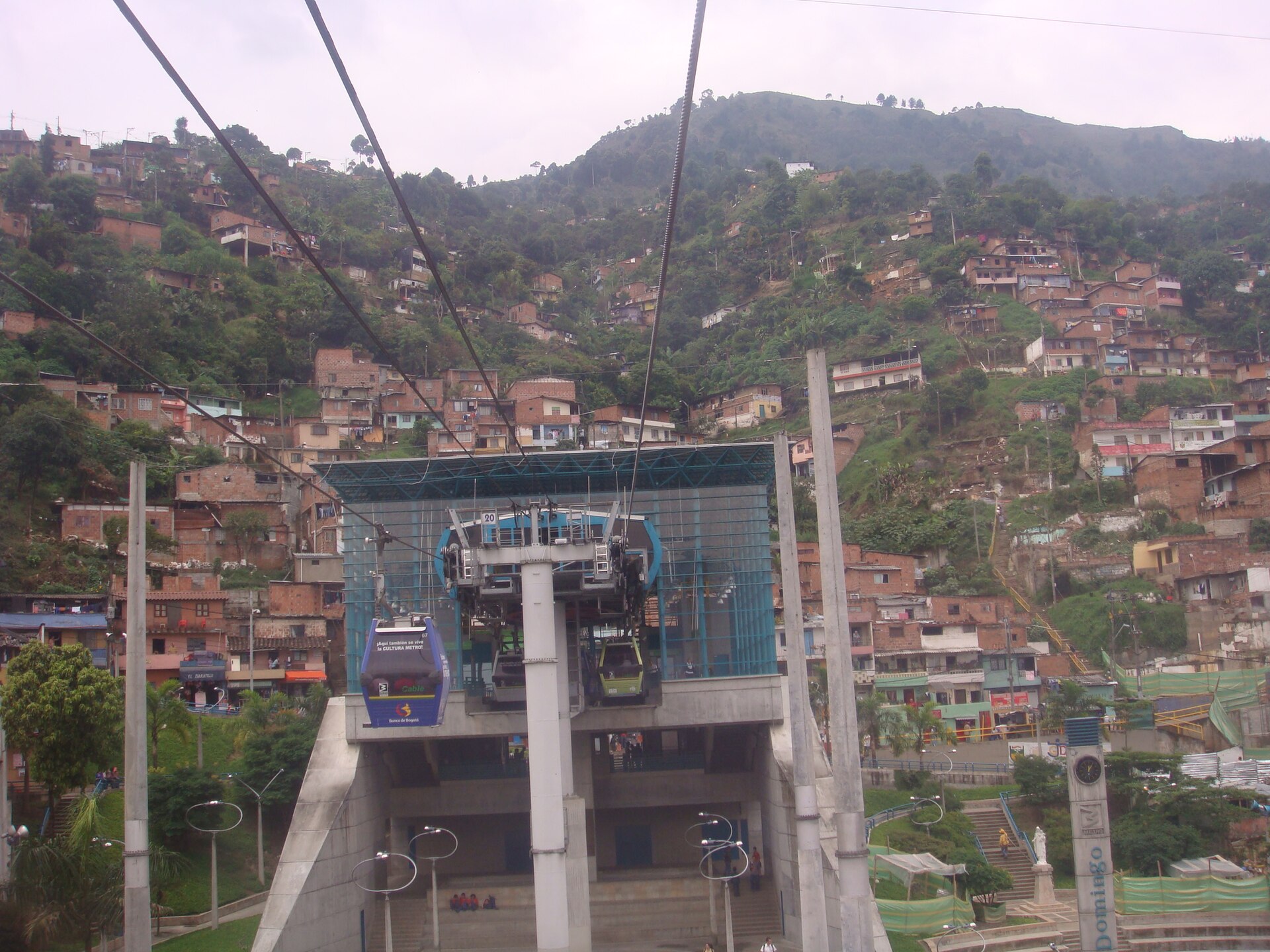Fotografía en color de una estación de teleférico con cabinas colgando de los cables, y detrás una ladera densamente construida con casas de ladrillo
