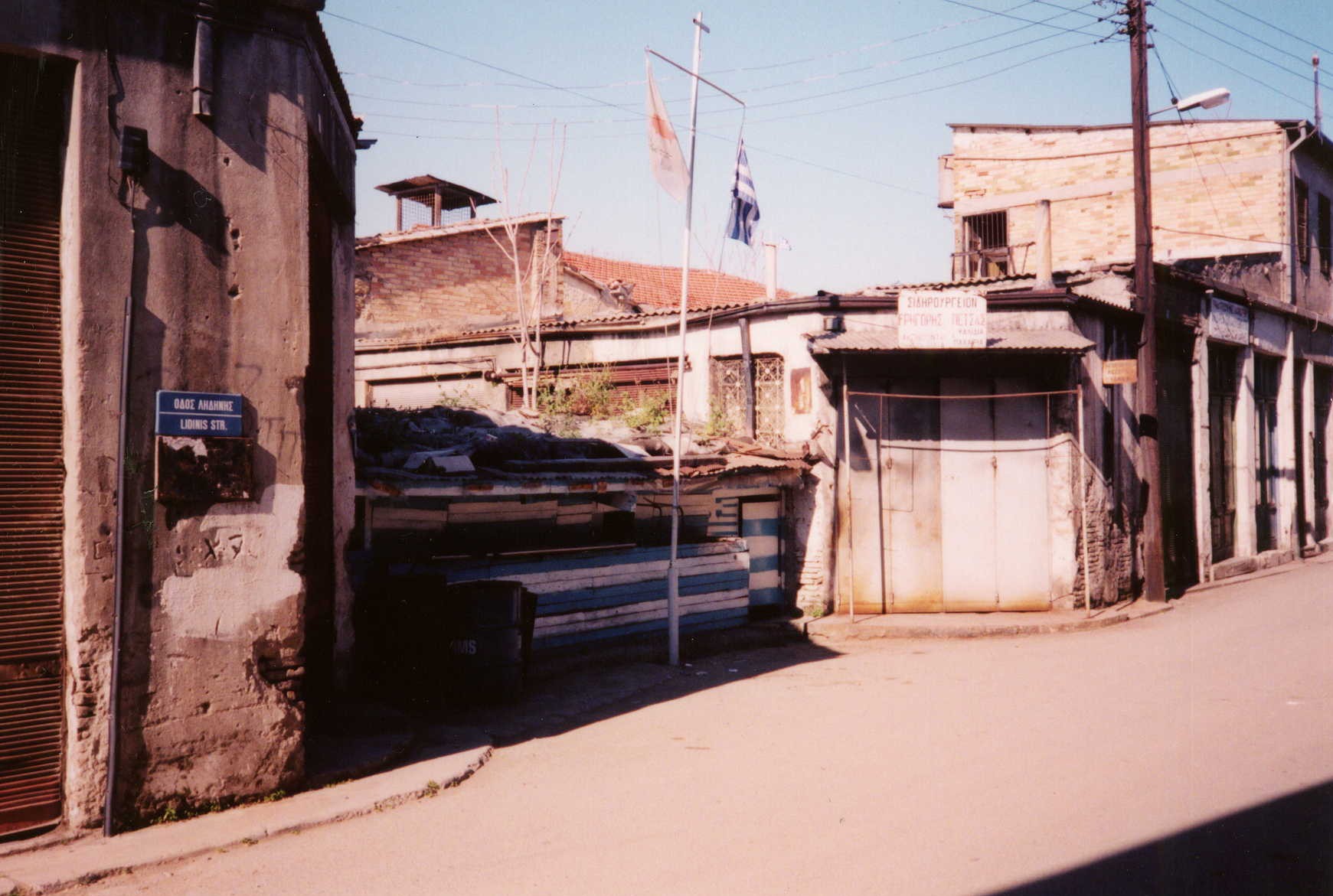 Fotografía en color de una calle estrecha cortada por una barricada de bidones pintados con rayas azules y blancas, con banderas de Grecia y de Chipre sobre un mástil