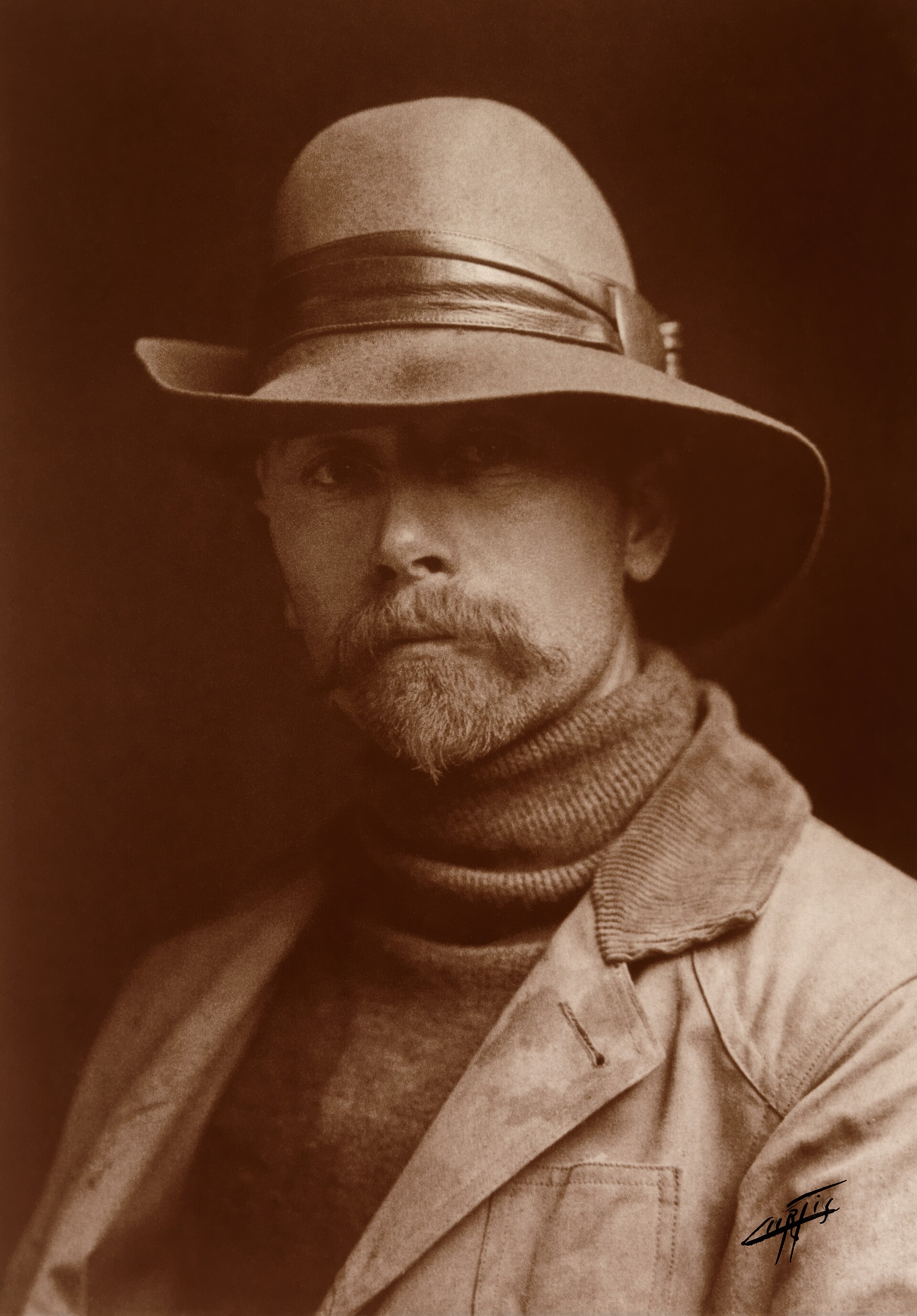 Autorretrato fotográfico de un hombre joven de perfil, con barba corta, chaqueta oscura y corbata, iluminado de lado sobre fondo neutro