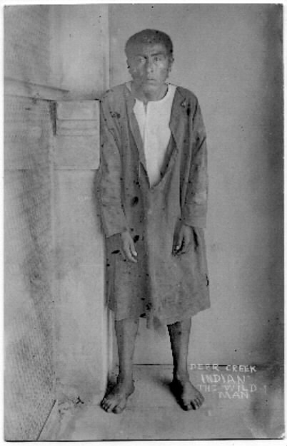 Fotografía de 1911 de Ishi de pie descalzo contra una pared, con una chaqueta de lona prestada; sobre la copia hay escrito a mano «Deer Creek Indian, the wild man»