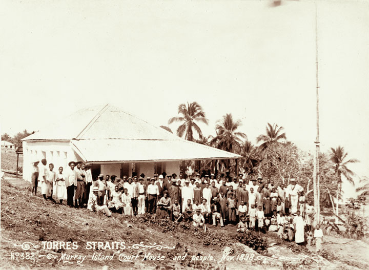Fotografía de 1898 de un edificio colonial de madera con tejado de chapa, con hombres meriam de pie y sentados delante