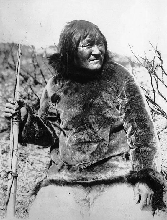 Retrato de un hombre mayor con parka de piel sentado al aire libre, sujetando el astil de un arpón, con matorrales al fondo
