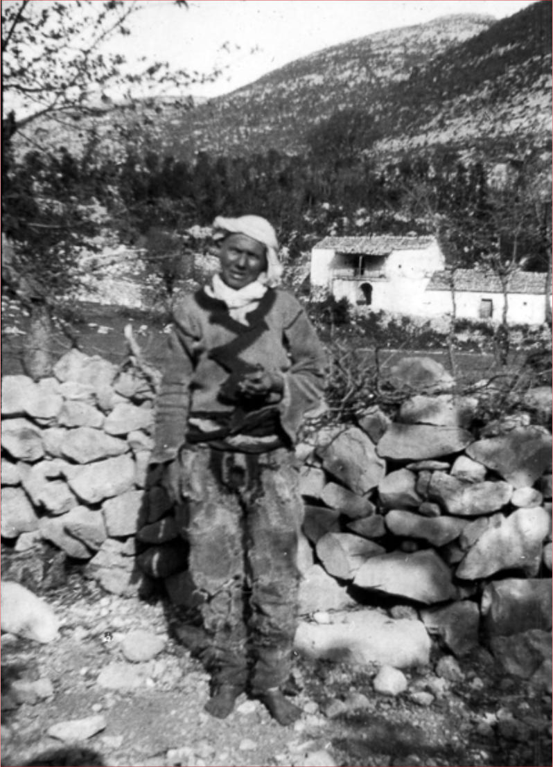 Fotografía de 1908 de una persona con ropa clara y cinturón, de pie en un terreno pedregoso de montaña