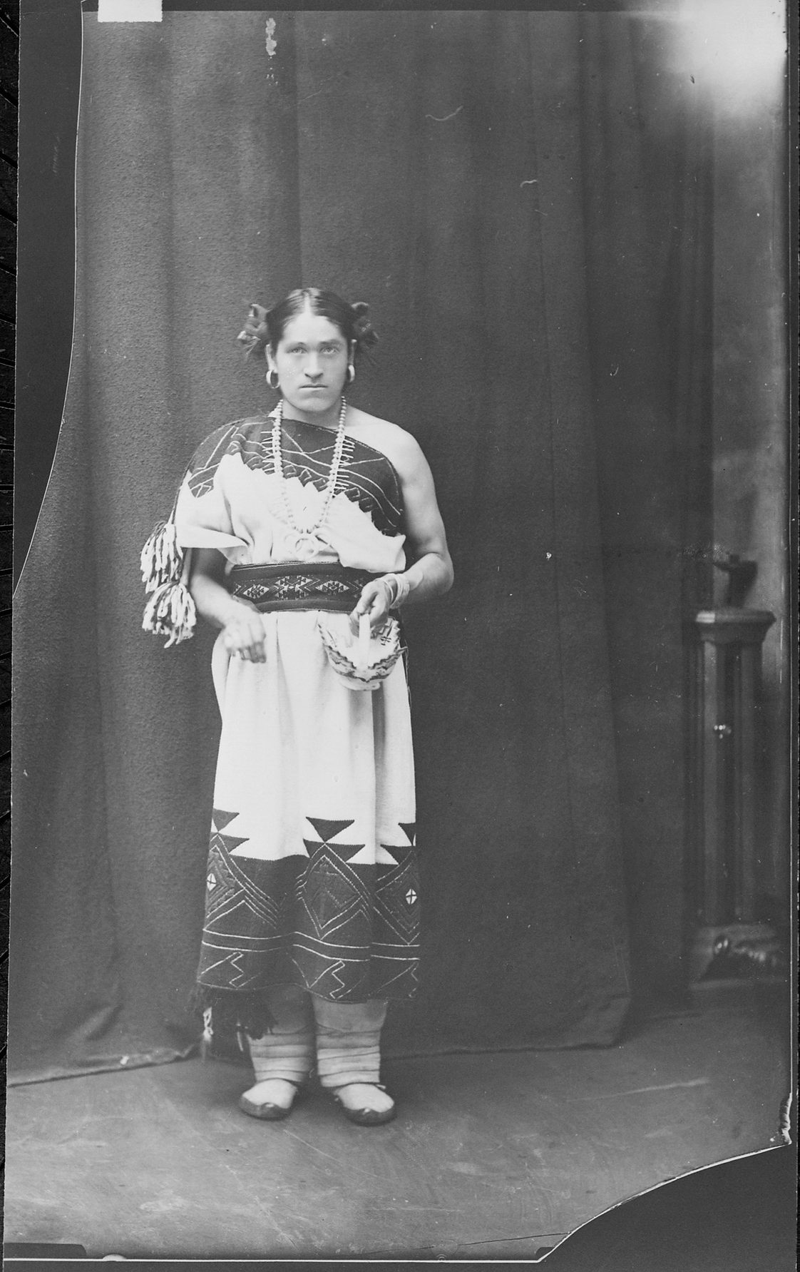 Retrato fotográfico de cuerpo entero de una persona zuñi con manta bordada, collares y el pelo recogido