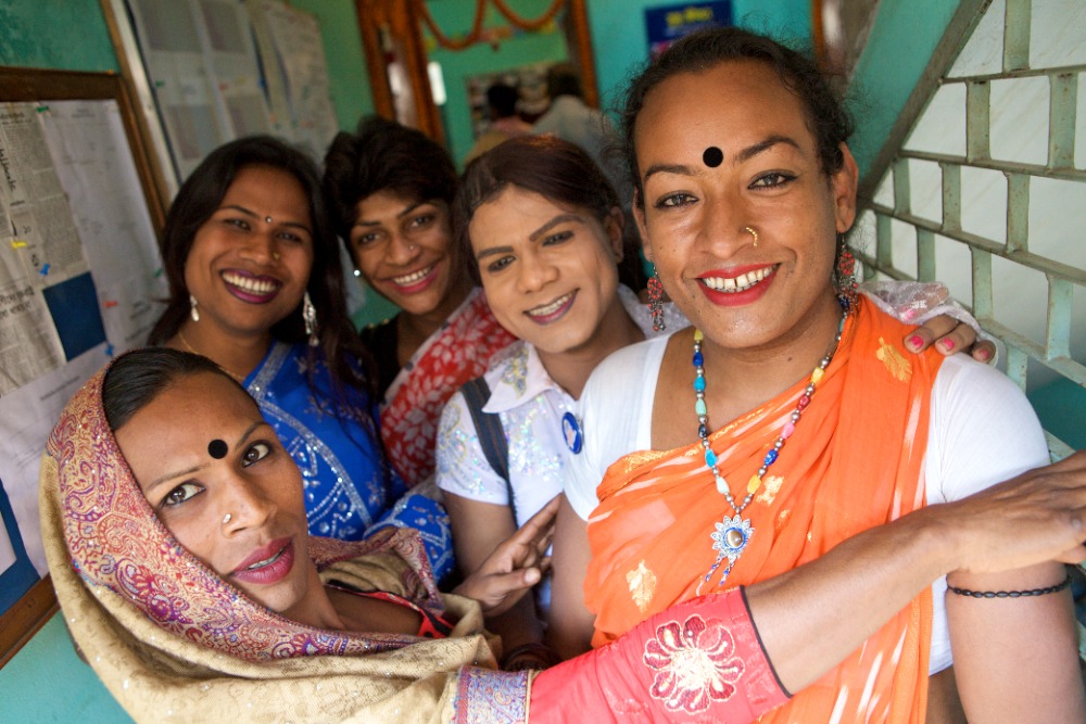 Fotografía en color de un grupo de personas sonrientes con saris de colores y bindi, reunidas en un interior