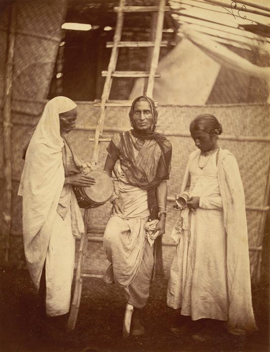 Fotografía del siglo XIX de tres personas en pie con saris y adornos, en un estudio fotográfico