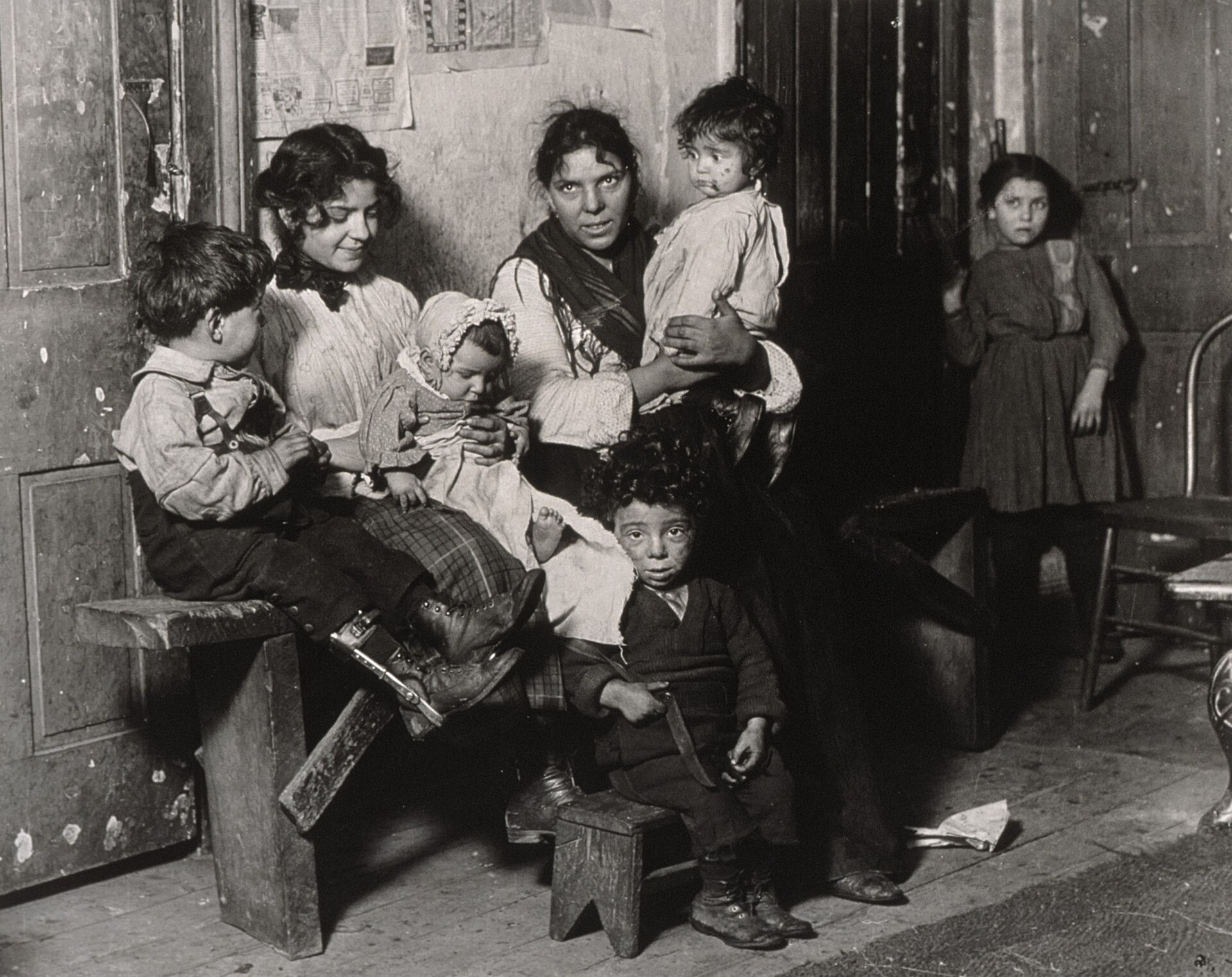 Fotografía antigua del interior de una vivienda humilde con una mujer, varios niños pequeños sentados en un banco y una niña de pie junto a la puerta