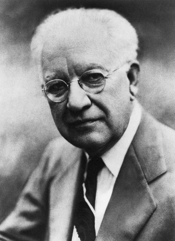 Retrato en blanco y negro de un hombre mayor con gafas redondas, pelo blanco, traje y corbata