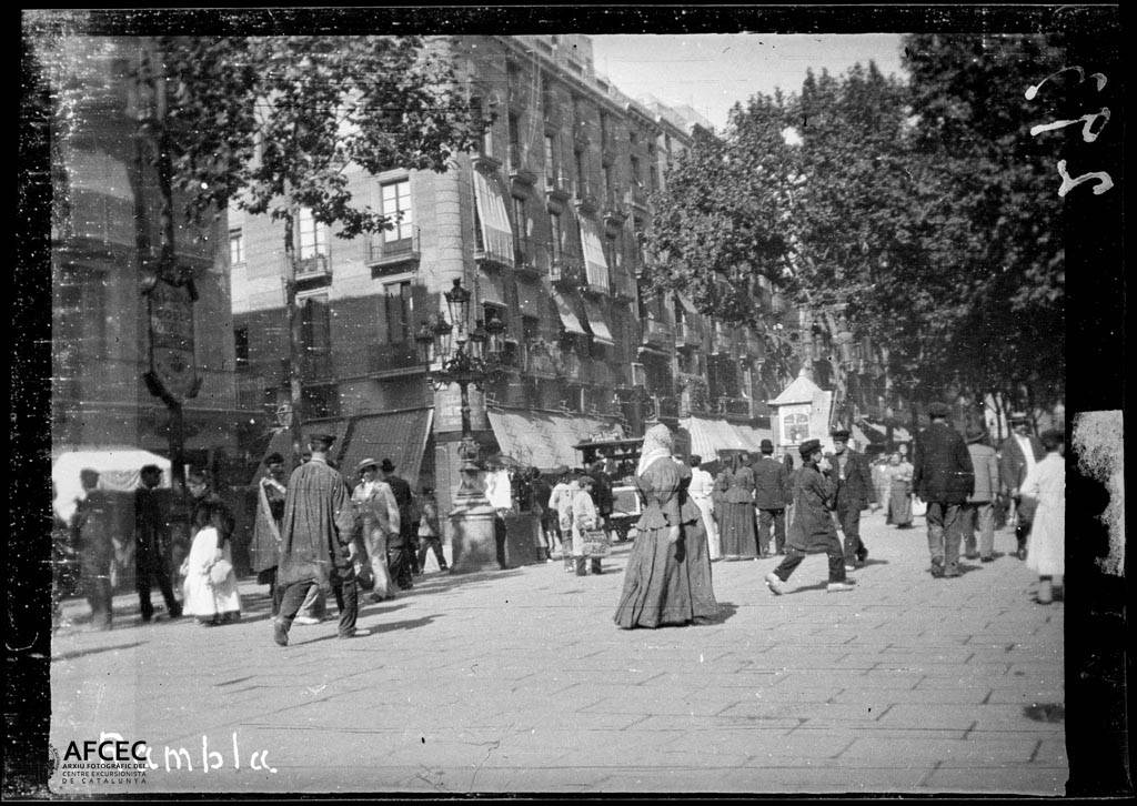 Fotografía antigua de un paseo arbolado con mucha gente caminando, mujeres con vestidos largos y hombres con sombrero, frente a edificios de varias plantas