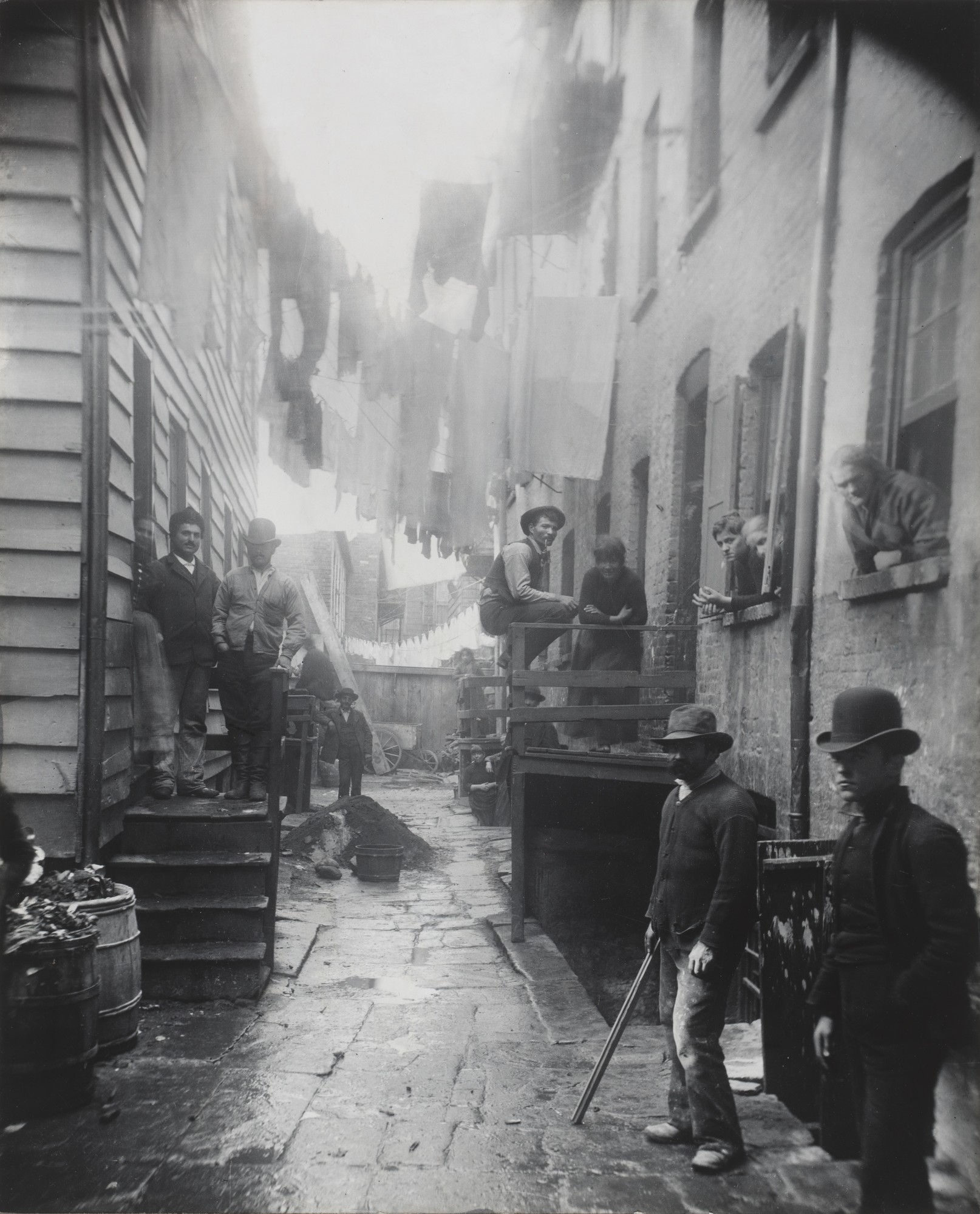 Fotografía antigua de un callejón estrecho entre edificios de ladrillo, con ropa tendida en lo alto y varios hombres con sombrero que miran a la cámara