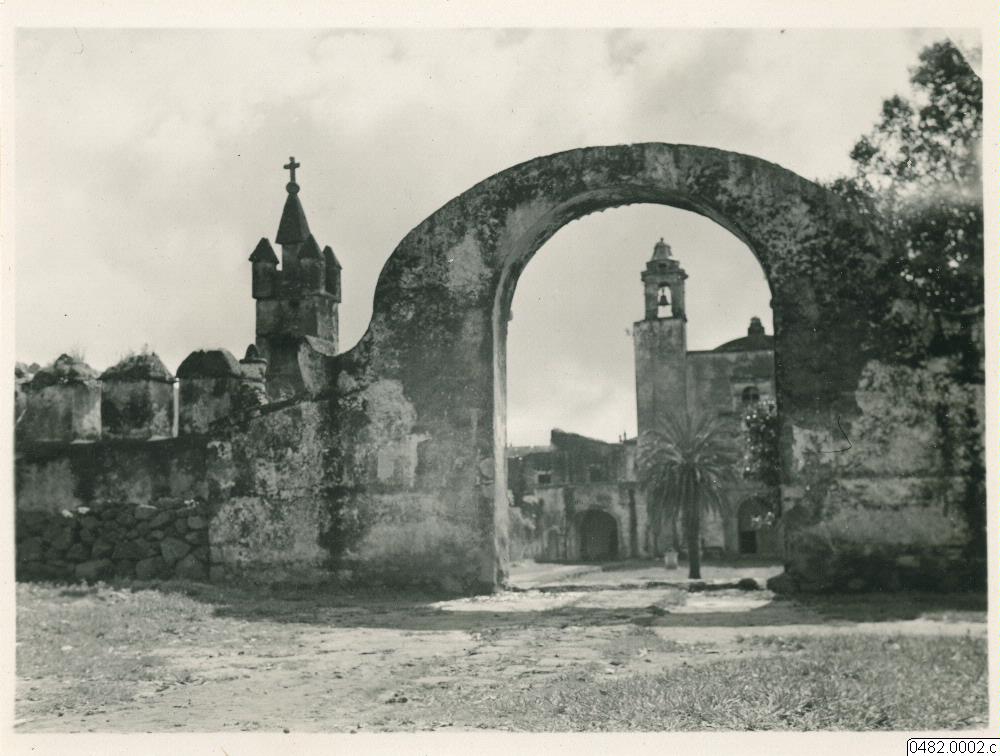 Fotografía antigua en sepia de un arco de piedra en ruinas que enmarca la torre de una iglesia colonial, con un muro bajo y un camino de tierra
