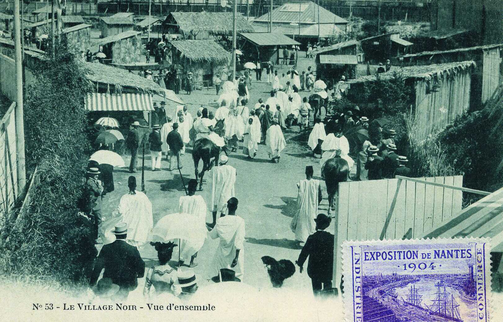 Postal de 1904 titulada «Le Village Noir — Vue d'ensemble» con calles de chozas, gente en túnicas blancas y visitantes franceses con sombreros de paja y parasoles