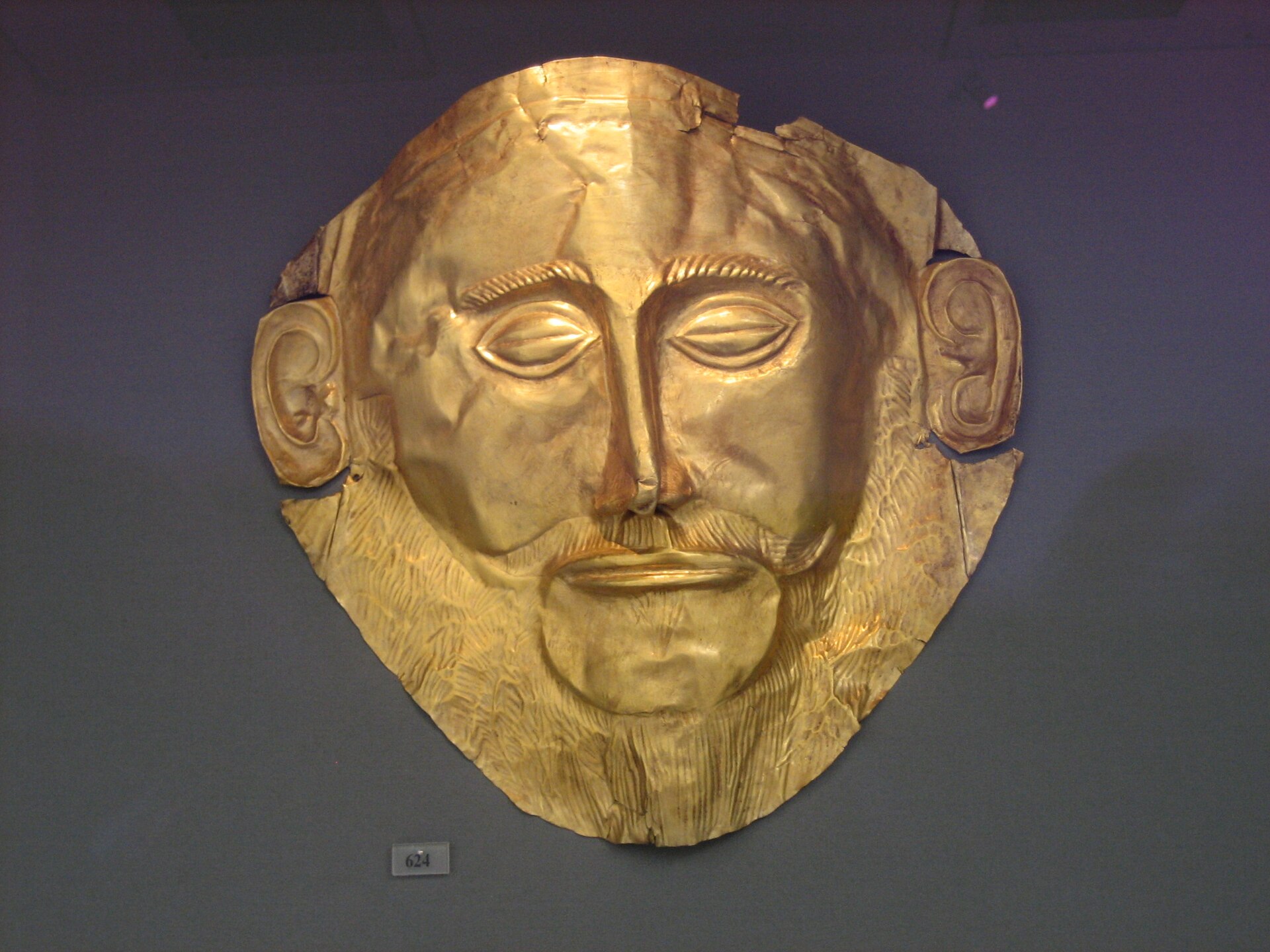 Máscara funeraria de oro batido con rasgos faciales estilizados, bigote y orejas marcadas