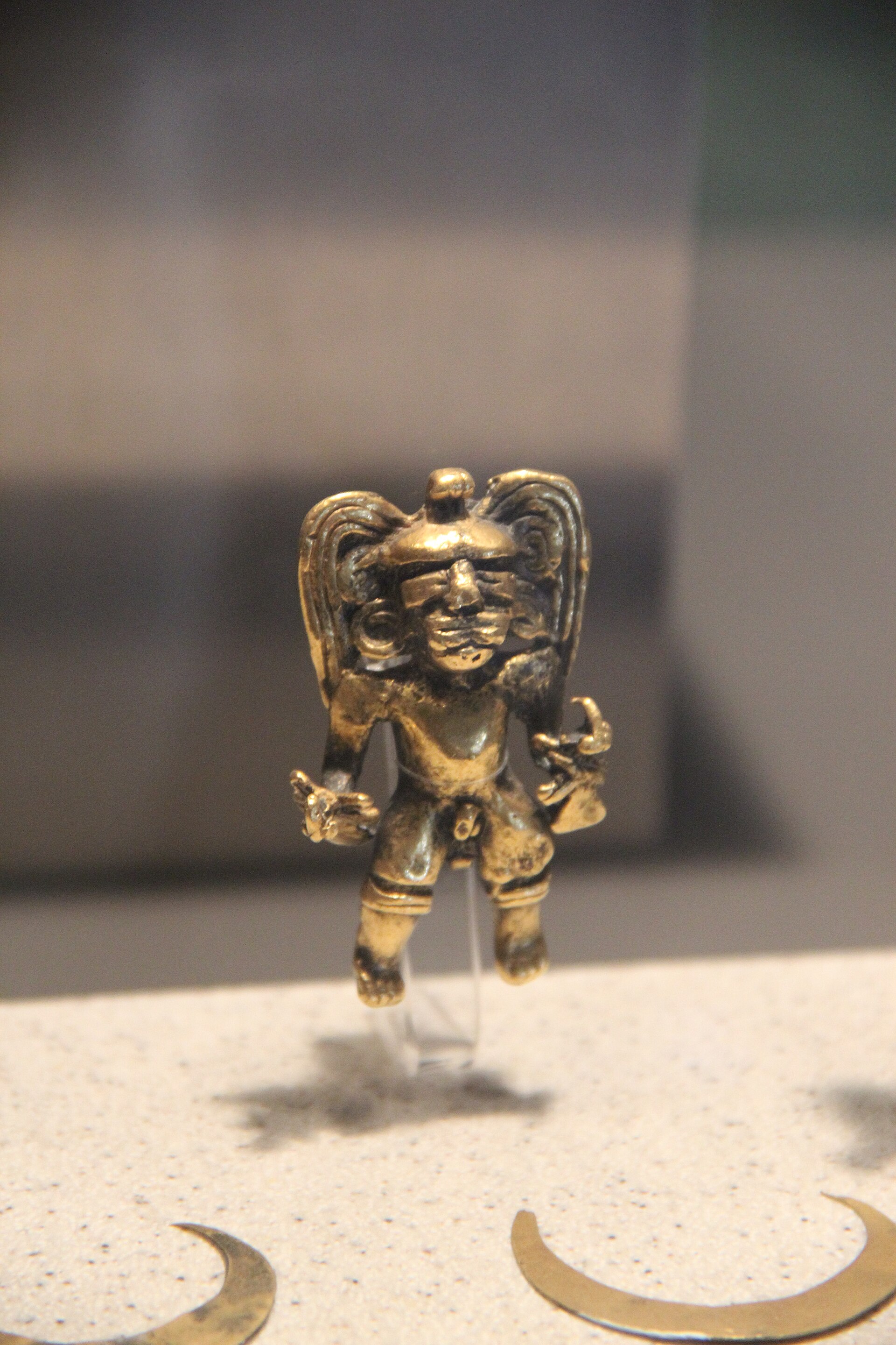 Pequeña figura humana de oro con tocado de alas, expuesta en una vitrina sobre fondo claro