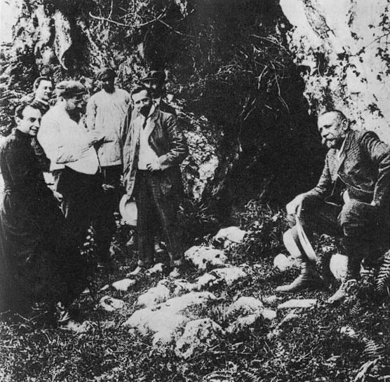 Fotografía de 1909 en la boca de una cueva con cuatro hombres: uno con sotana, otro sentado a la derecha y dos de pie al fondo