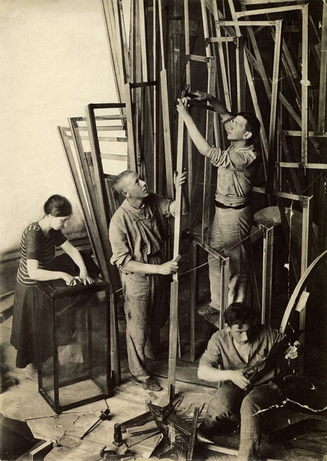 Fotografía en blanco y negro de un hombre subido a una escalera junto a una gran maqueta de madera en espiral, con varios ayudantes trabajando en el taller
