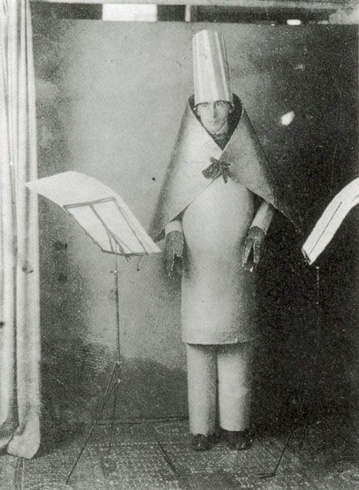 Fotografía en blanco y negro de un hombre dentro de un rígido traje de cartón con forma de columna, una capa angulosa sobre los hombros y un sombrero alto de chamán