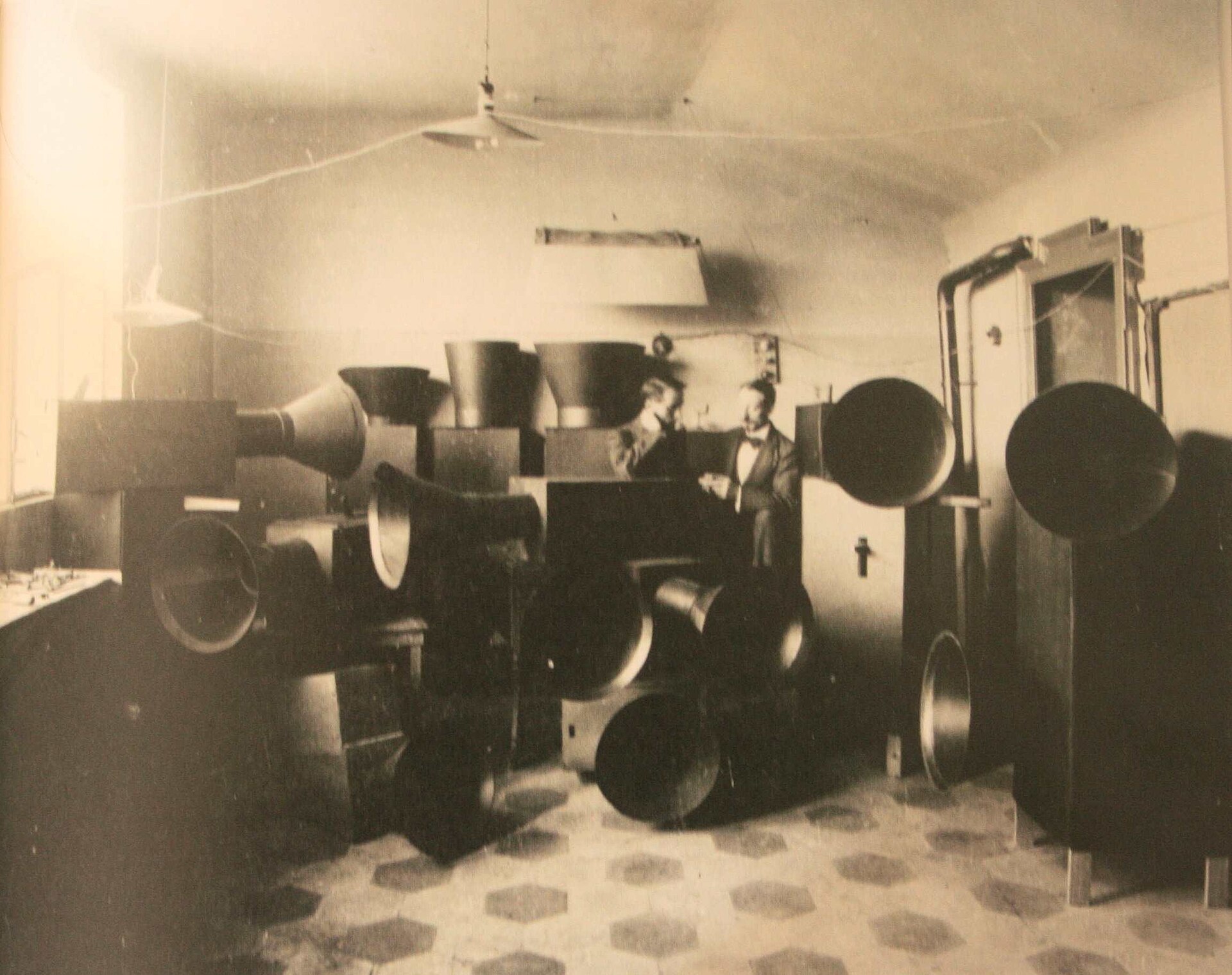Fotografía de un taller con dos hombres junto a una fila de grandes cajas de madera con bocinas metálicas, los instrumentos de ruido futuristas
