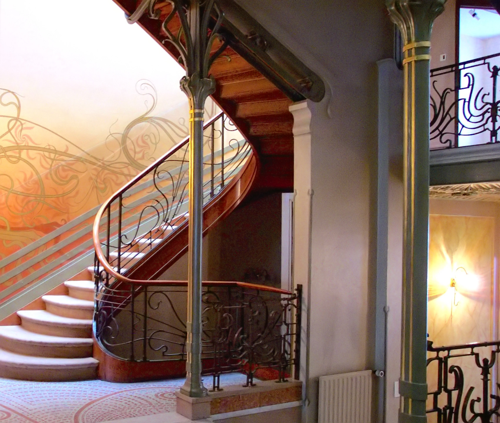 Interior de una casa con una escalera curva de barandilla de hierro, una columna metálica cuyo capitel se abre en tallos y paredes con motivos lineales ondulados
