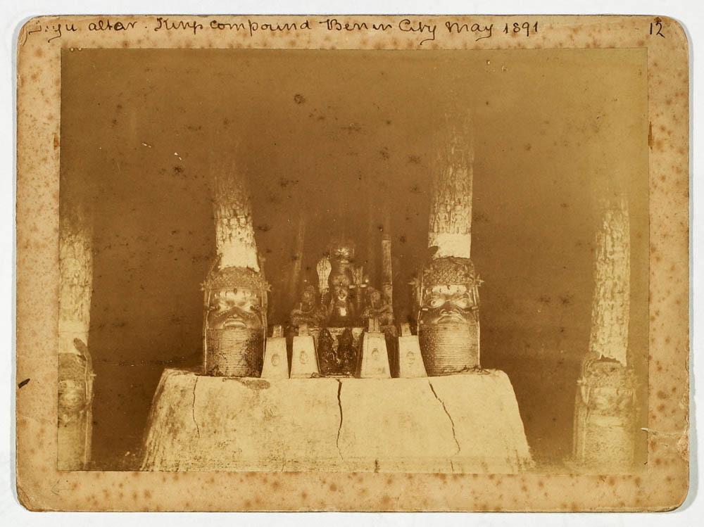 Fotografía antigua de un altar de barro con colmillos de marfil tallados sostenidos sobre cabezas de latón, dentro de un recinto con pilares, con anotación manuscrita arriba