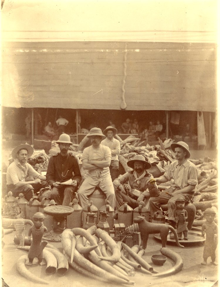 Fotografía de 1897 con seis europeos con salacot sentados entre colmillos de marfil, cabezas de latón y figuras amontonadas en el suelo