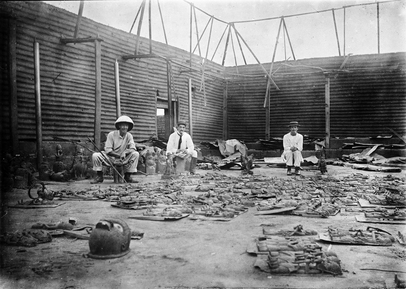 Interior de un recinto incendiado con las paredes de tablones en pie, el suelo cubierto de escombros y objetos, y tres europeos con salacot sentados entre ellos