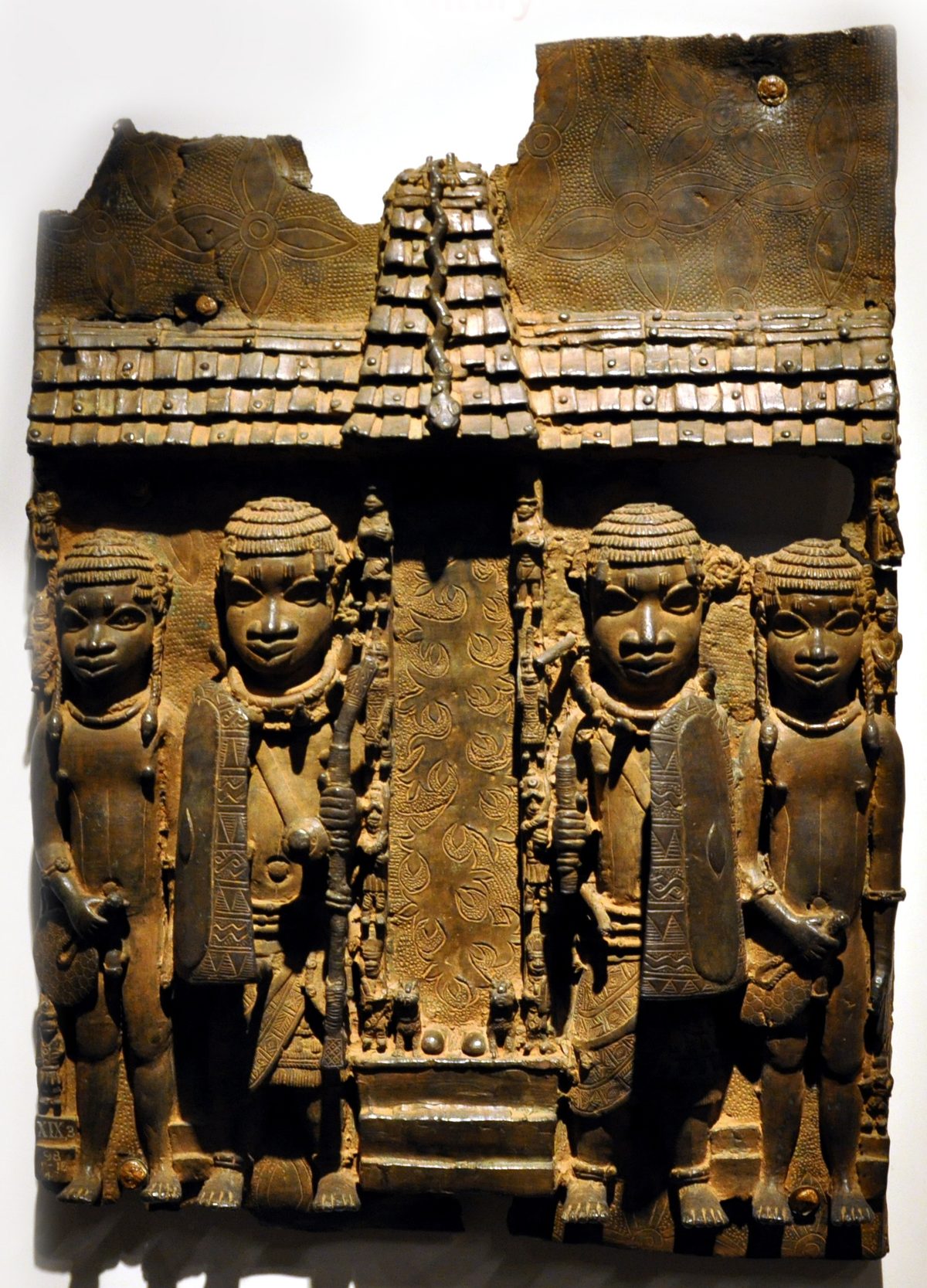Placa rectangular de latón con cuatro figuras en relieve bajo un tejado de palacio, con collares de coral, faldellines y armas
