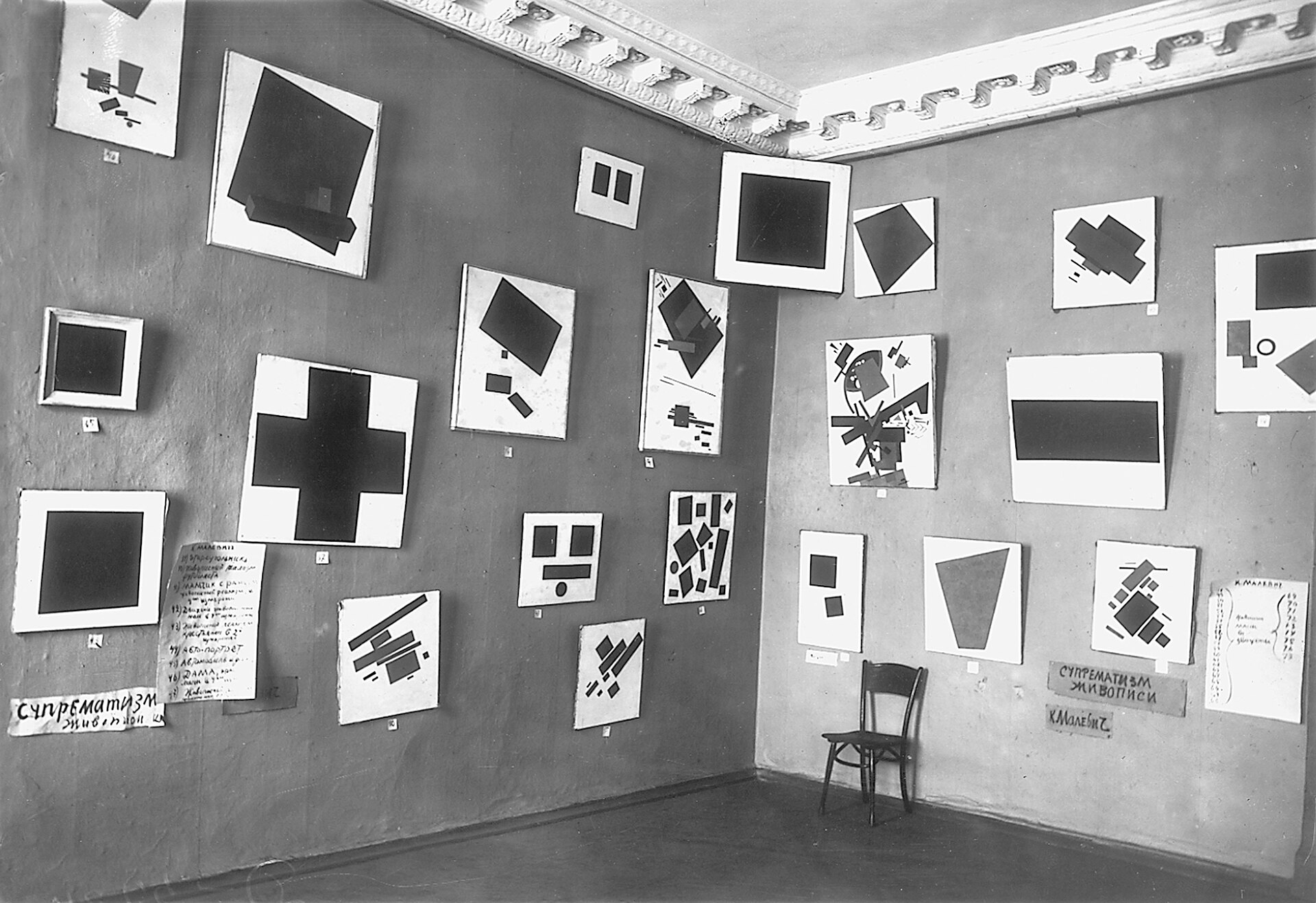 Fotografía en blanco y negro de una esquina de una sala de exposición con muchas pinturas geométricas colgadas y un cuadrado negro arriba en el rincón