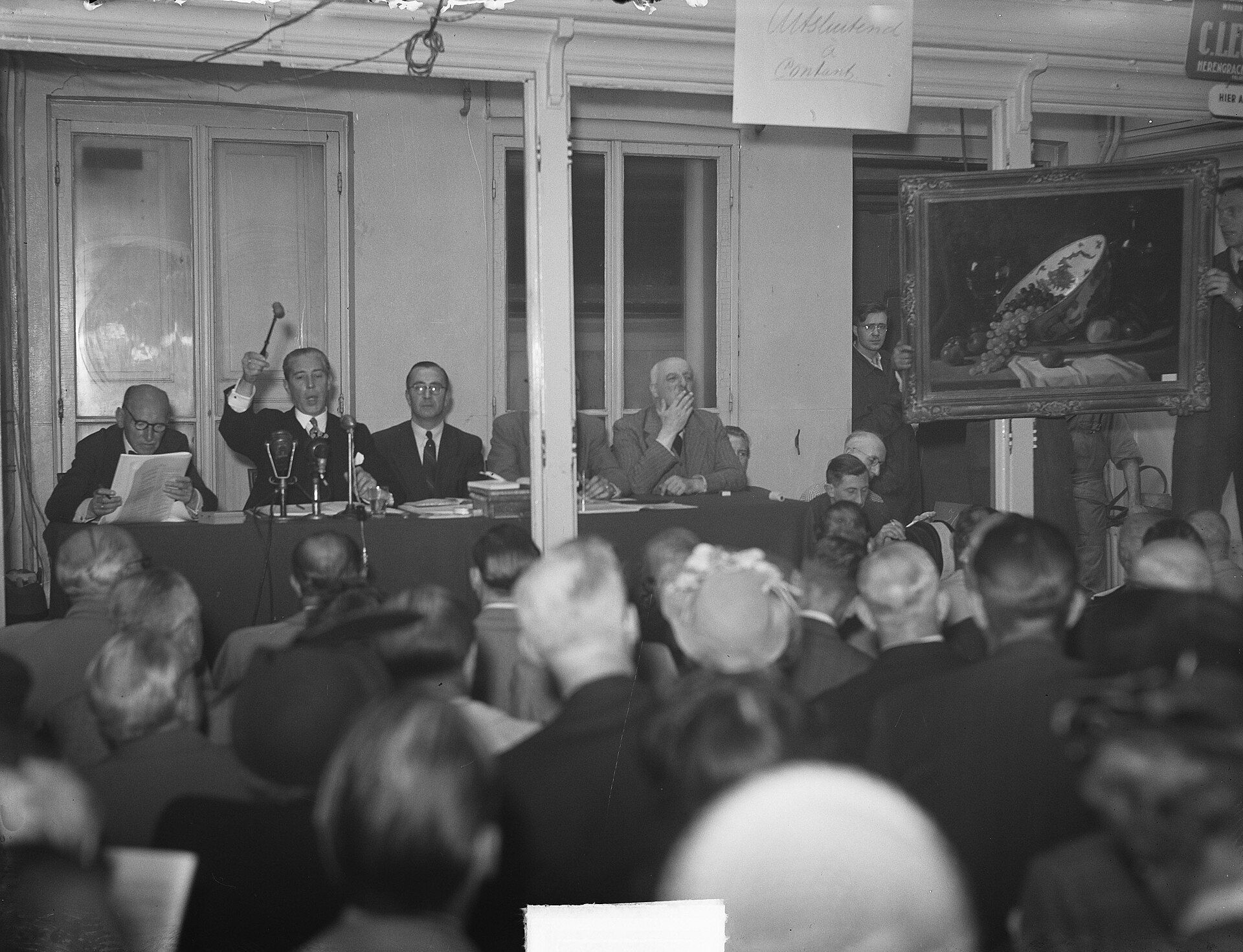 Sala llena de público en la subasta de los bienes de Van Meegeren en 1950, con un cuadro expuesto y varias manos levantadas pujando