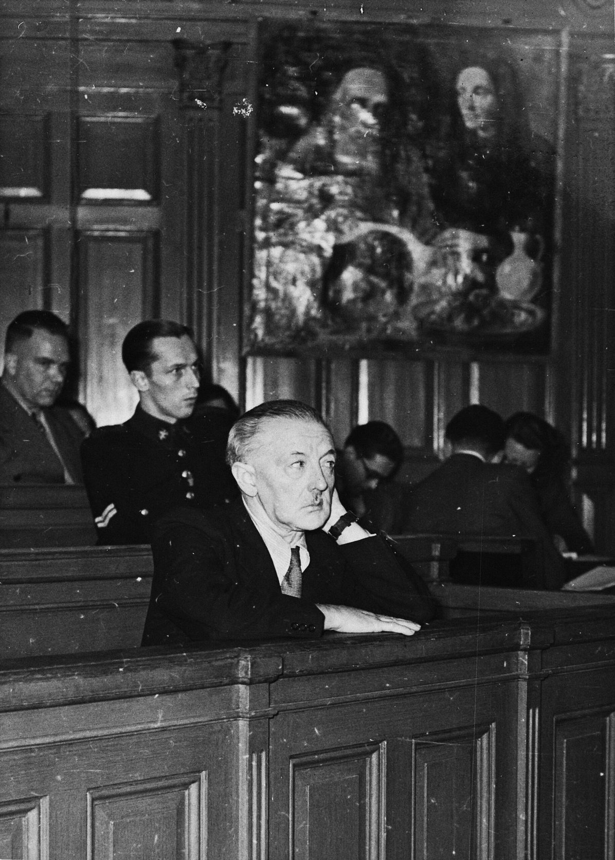 Van Meegeren sentado en el banquillo durante su juicio de 1947, con varios cuadros colgados en la pared del fondo de la sala