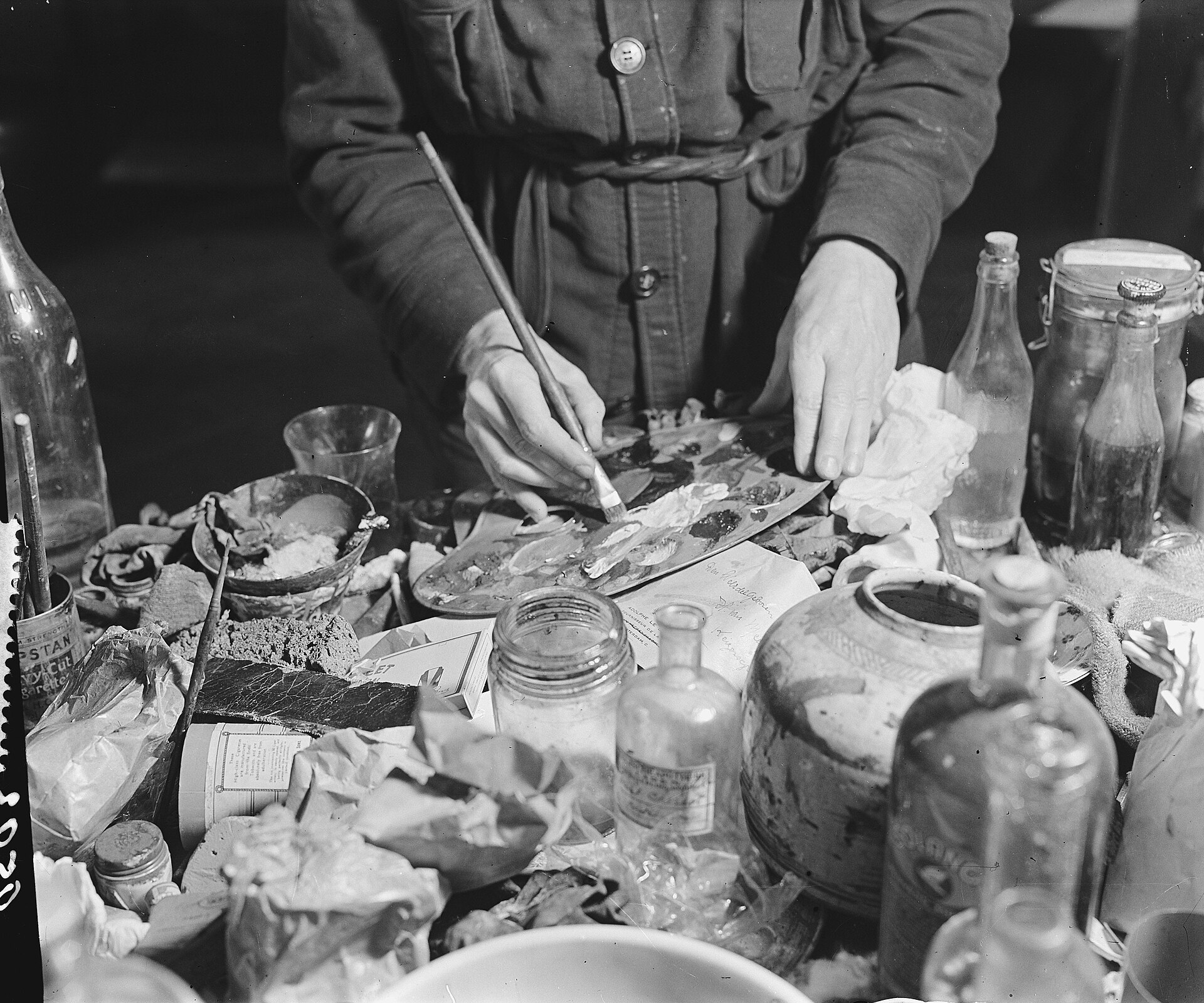 Fotografía en blanco y negro de unas manos que pintan con un pincel fino junto a una mesa llena de frascos, botellas y trapos