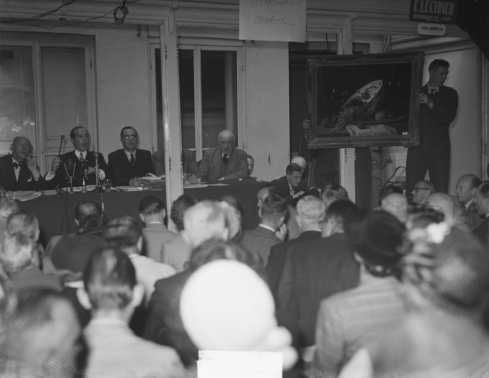 Fotografía en blanco y negro de una sala de subastas llena de público sentado, con cuadros expuestos al fondo