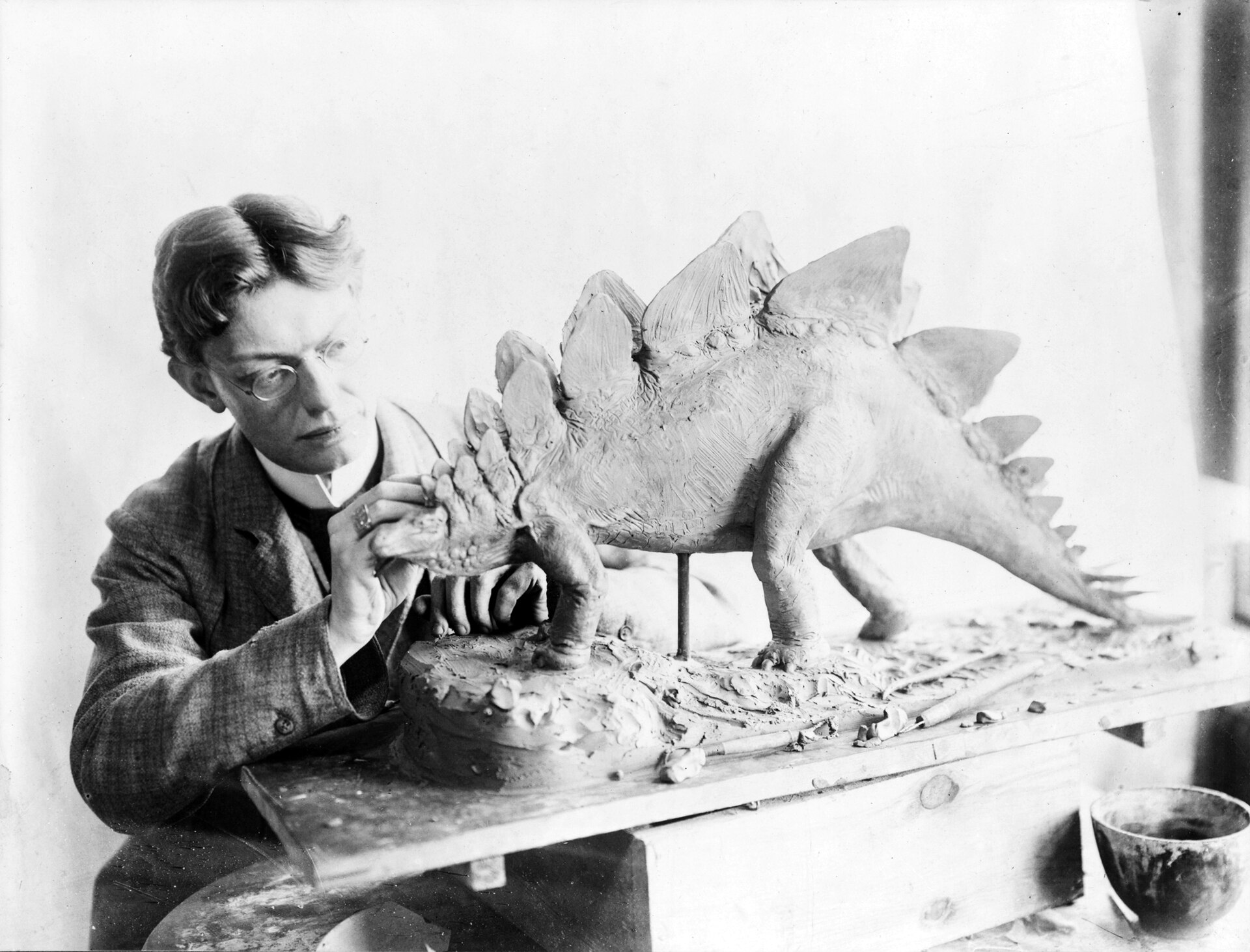 Fotografía en blanco y negro de un hombre trabajando en un modelo de un dinosaurio con placas en el lomo, en un taller