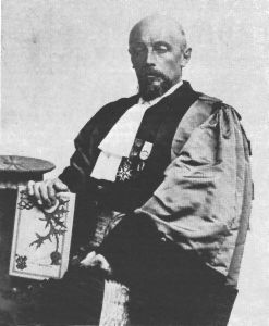 Retrato fotográfico antiguo de un hombre calvo con barba canosa, vestido con toga académica con condecoraciones, que sostiene un documento enmarcado