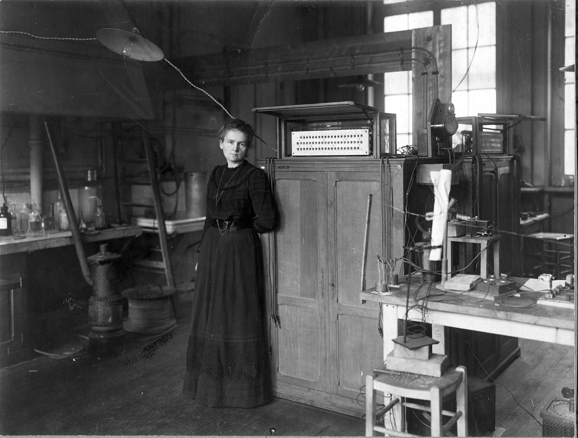 Marie Curie, de pie, con bata oscura, ante una mesa de laboratorio llena de matraces y aparatos de vidrio