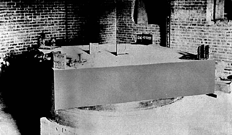 Fotografía antigua de un gran bloque cuadrado de piedra sobre un soporte, con pequeños espejos y piezas ópticas montados en las esquinas