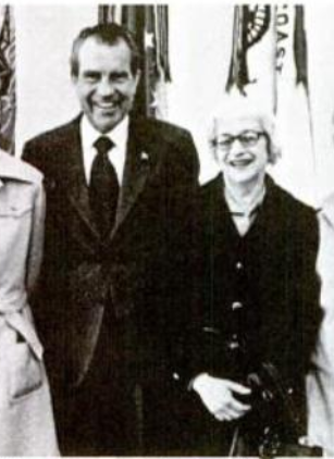 Fotografía en blanco y negro de una mujer de pelo corto y gafas, sonriente, con abrigo oscuro, junto a un hombre trajeado que sonríe, en un despacho con banderas al fondo