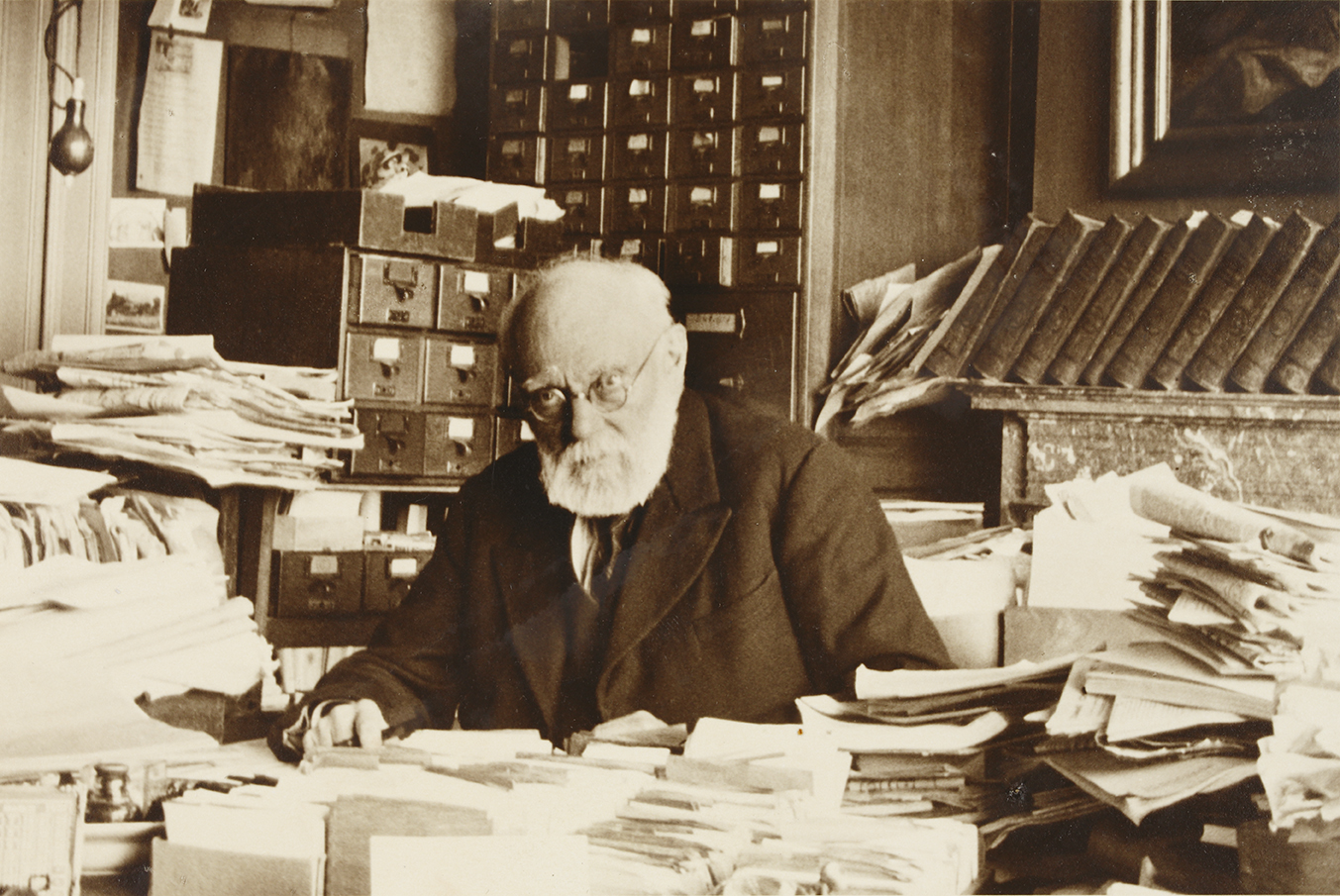 Fotografía en sepia de un hombre mayor con barba blanca sentado ante un escritorio cubierto de papeles apilados, con estanterías y archivadores de cajones detrás