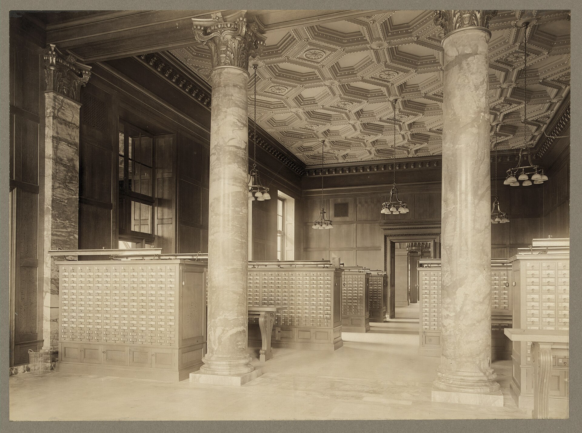 Fotografía antigua de una sala de biblioteca con columnas corintias y techo de casetones, ocupada por muebles bajos de madera con cientos de cajones de fichas