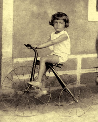 Fotografía antigua en sepia de un niño de unos diez años con el pelo corto y ropa clara, sentado en un triciclo de ruedas grandes junto a un muro