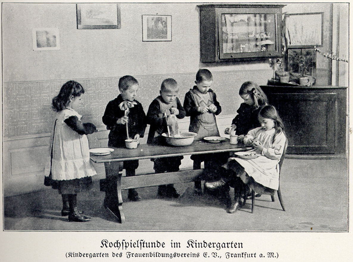 Fotografía antigua de un grupo de niños y niñas pequeños con delantales, de pie alrededor de una mesa de madera, jugando con cuencos y utensilios de cocina