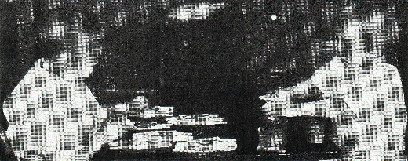 Fotografía antigua en blanco y negro de dos niños pequeños sentados a una mesa baja, manipulando piezas y tablillas de madera