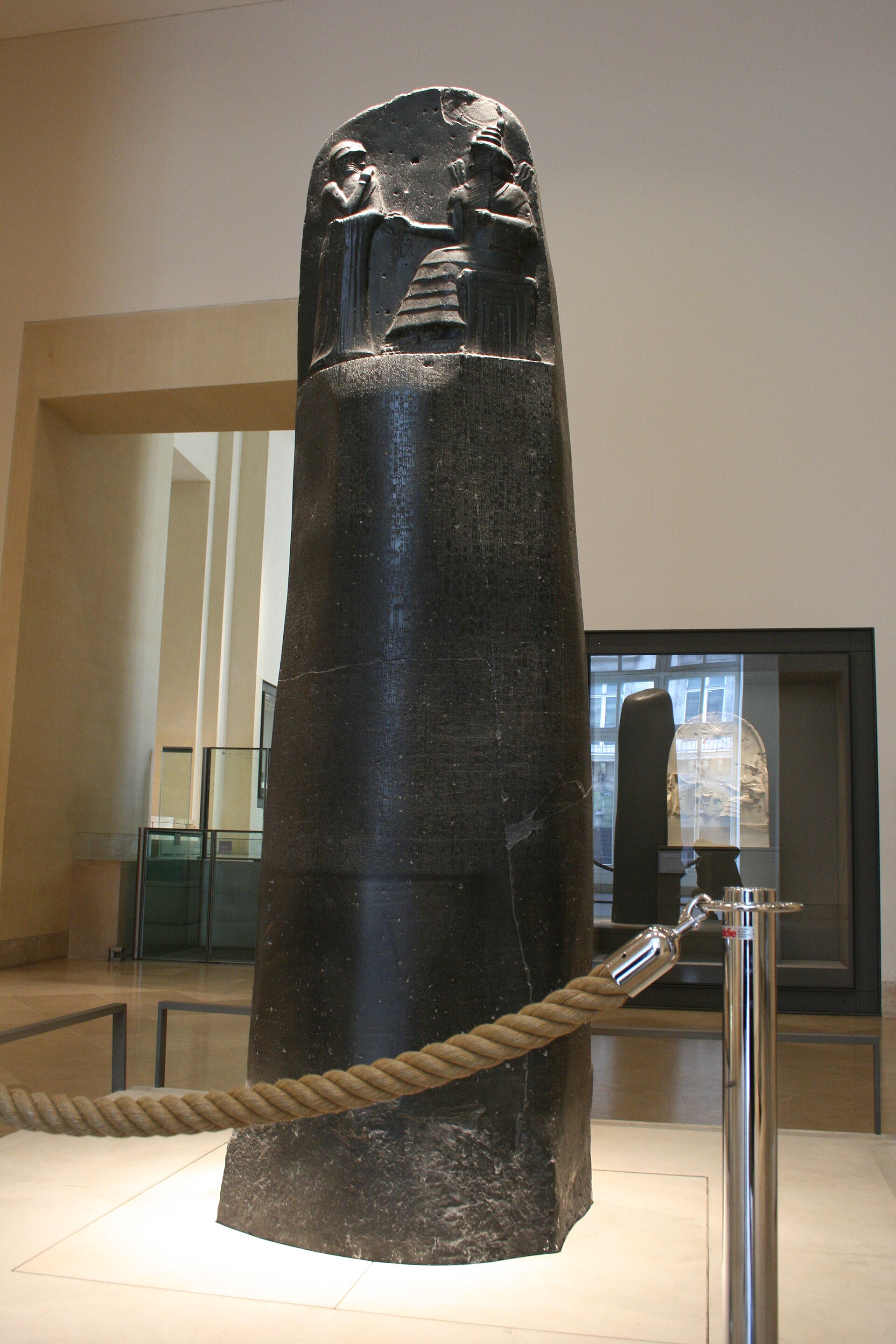 Estela de piedra negra de más de dos metros, con el relieve arriba y el resto cubierto de escritura cuneiforme, expuesta en una sala de museo detrás de un cordón