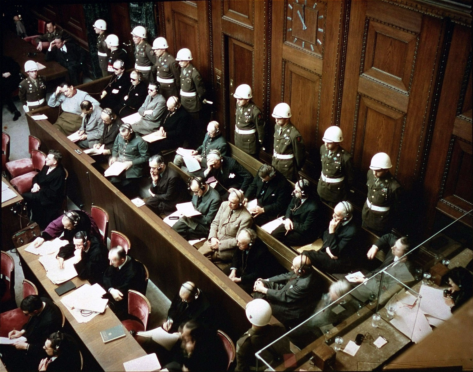 Fotografía en color tomada desde arriba del banquillo de los acusados, con dos filas de hombres de traje sentados y detrás una fila de policías militares con casco blanco