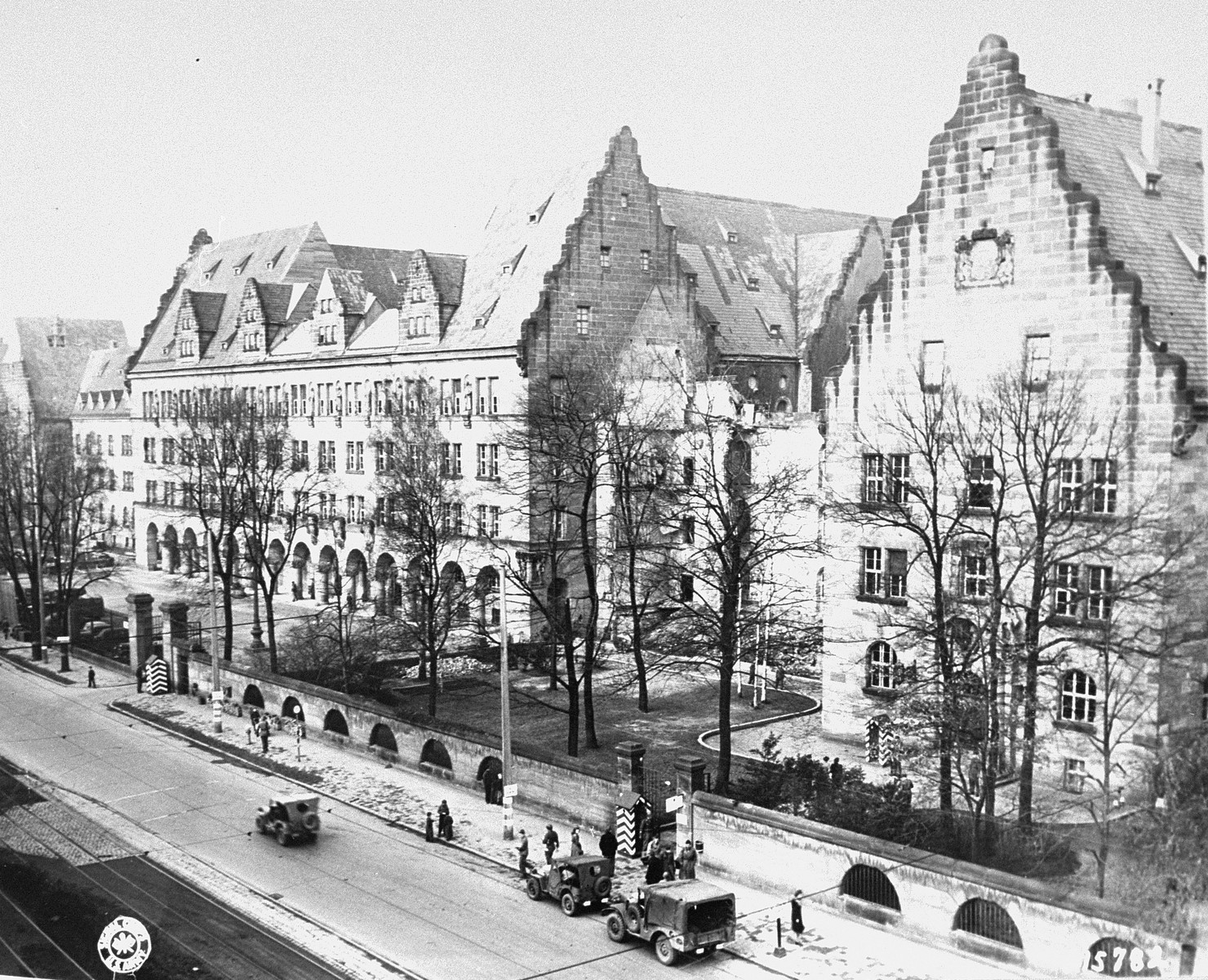 Fotografía aérea en blanco y negro de un gran edificio de piedra con tejados escalonados, rodeado de árboles sin hojas y con vehículos militares en la calle