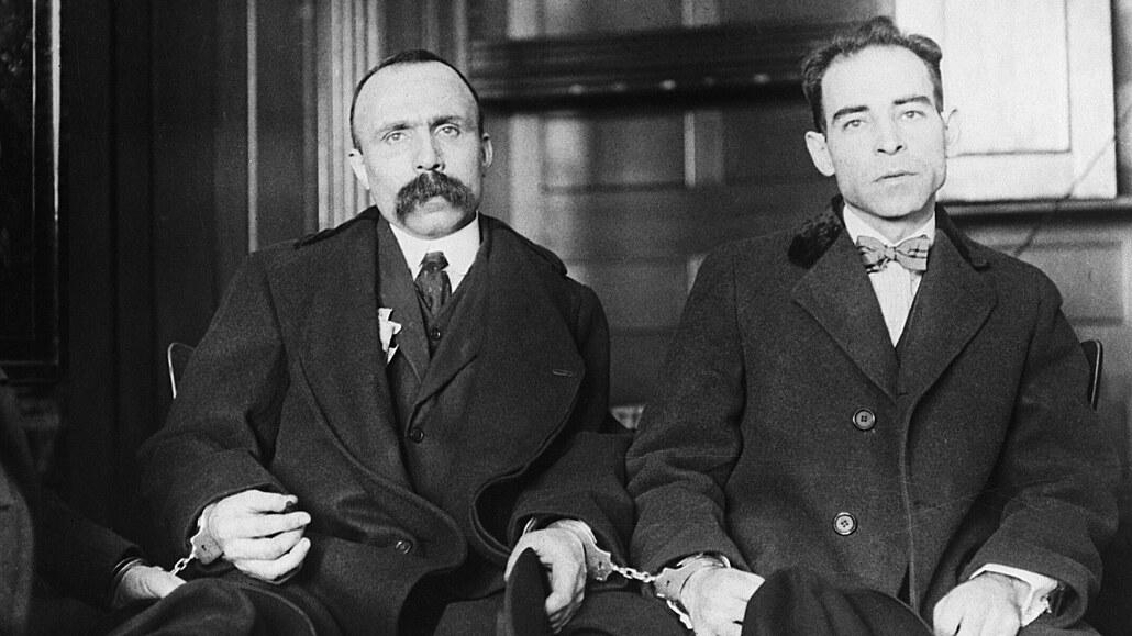 Fotografía en blanco y negro de dos hombres sentados, trajeados y esposados entre sí; el de la izquierda con un gran bigote y el de la derecha más delgado y con pajarita