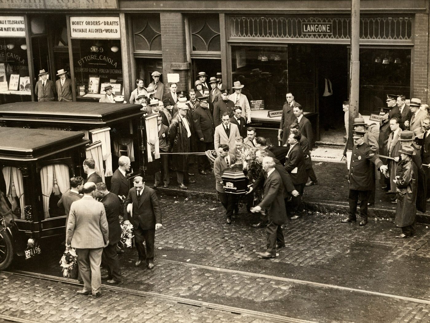 Fotografía de una calle con una multitud y varios hombres que sacan un féretro de un local con un rótulo de funeraria hacia un coche fúnebre